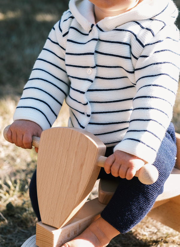 La Manufacture De Layette – Abrigo de Punto Les Basiques - Écru Rayas Marines, bebé con chaqueta de rayas blancas y azul marino sobre un juguete de madera