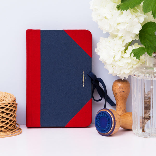 Ines De La Fressange – Carnet Médium Ines - Bleu, carnet de notes bleu et rouge posé sur une table avec des fleurs blanches