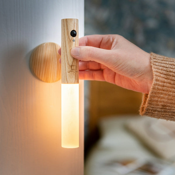 Gingko Design – Leuchte Smart Baton – Weiße Esche, eine Hand hält eine eingeschaltete Lampe aus hellem Holz und Opalglas