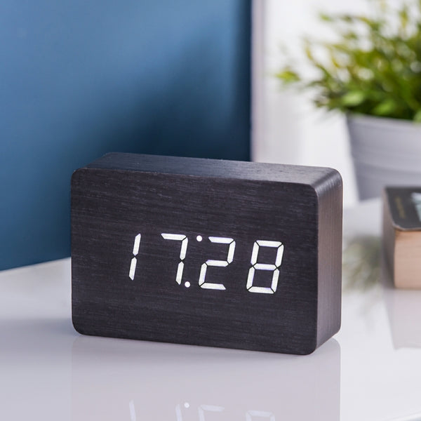Gingko Design – Wecker Brick Click - Schwarz / Weiße LED, Digitalwecker aus schwarzem Holz mit Zeitanzeige auf einem weißen Tisch